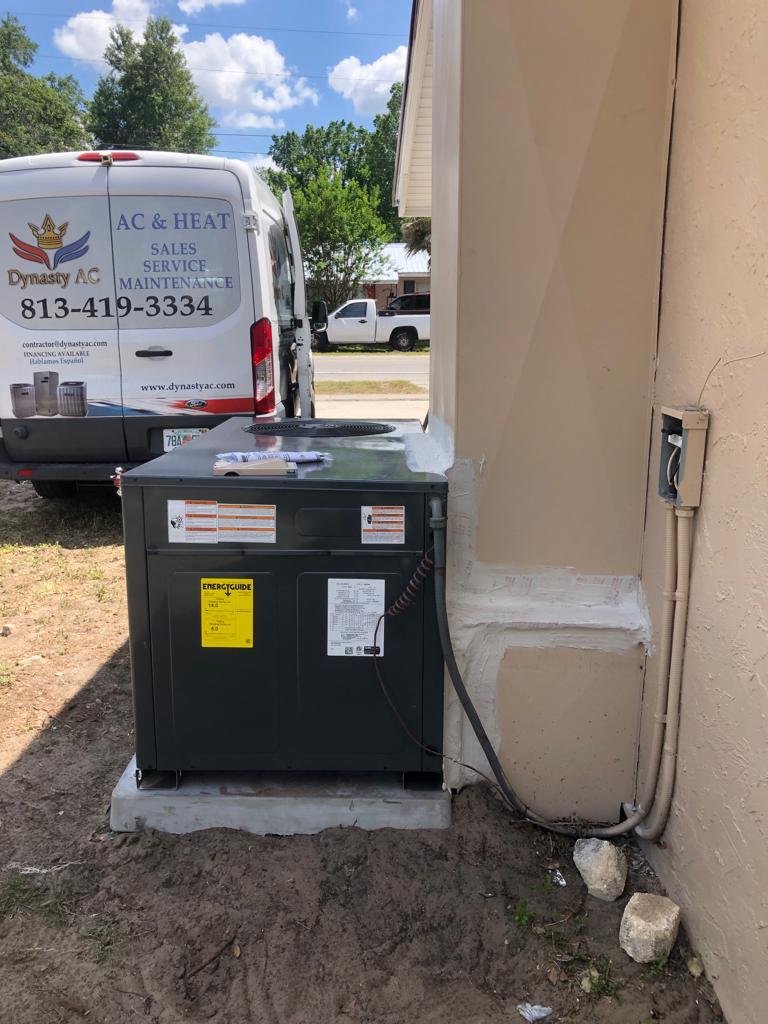 New outdoor AC condenser installation beside a house with a Dynasty AC service van parked behind it