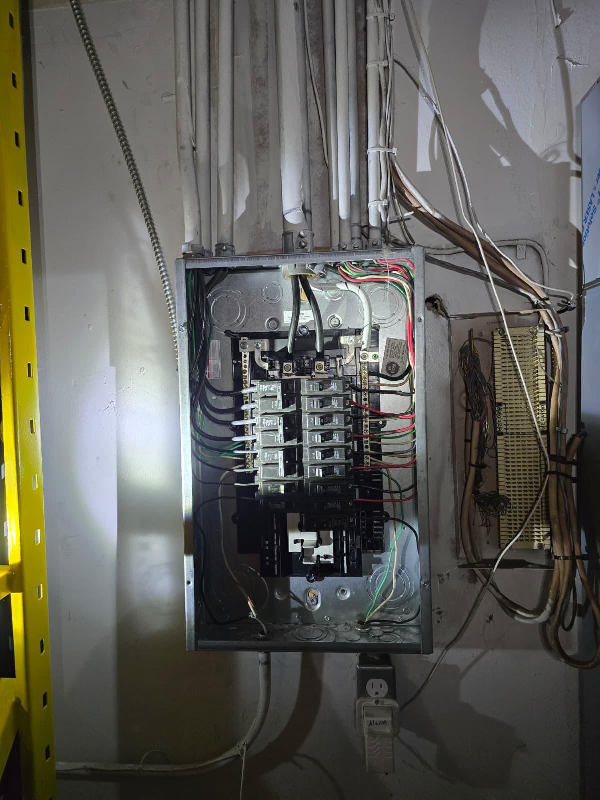 Open indoor electrical panel with breakers and wiring beside a patch panel